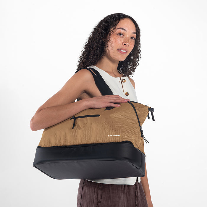 A woman wearing a Sherpani Boston Sand Anti-theft Weekender against a white background. #color_sand-tan