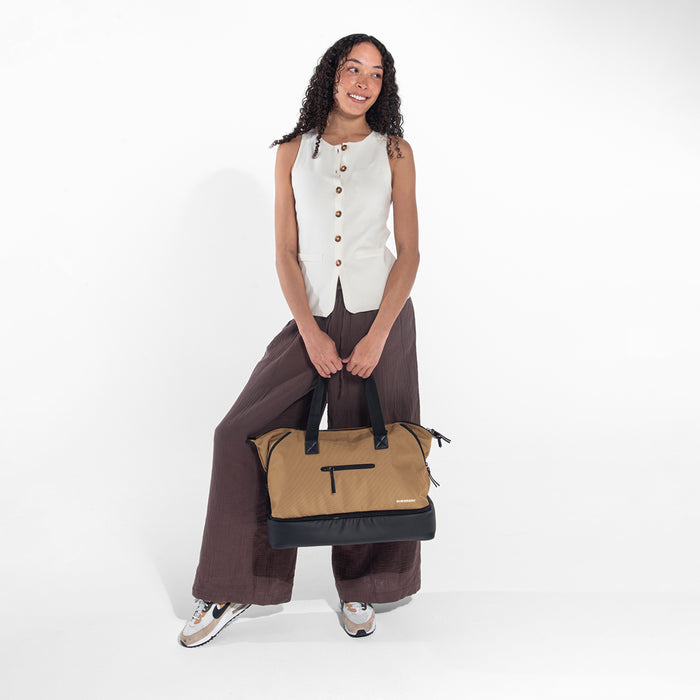 A woman holding a Sherpani Boston Sand Anti-theft Weekender against a white background. #color_sand-tan