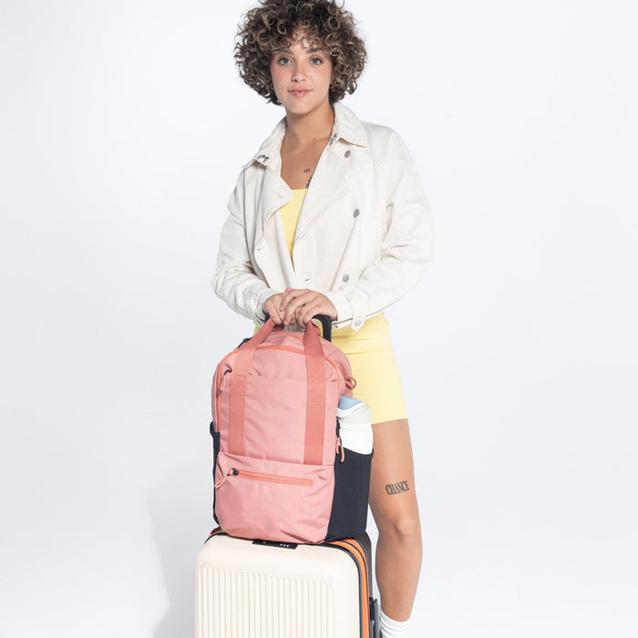 Woman holding a Sherpani Camden Desert Pink Backpack on top Sherpani Meridian Chai Beige luggage on white background. #color_desert-pink