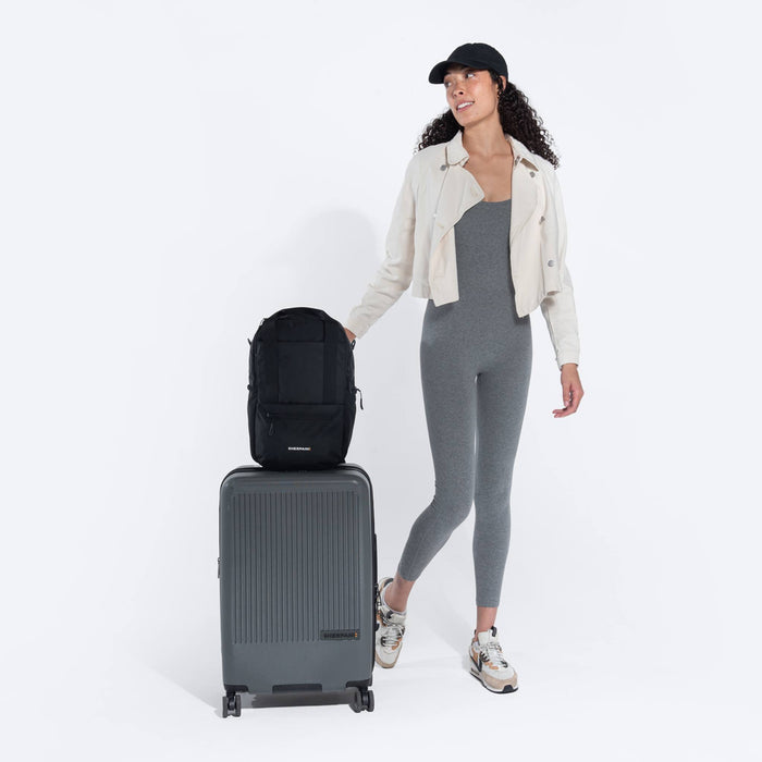 Woman holding a Sherpani Camden Raven Black Backpack on top Sherpani Meridian Granite luggage on white background. #color_raven-black