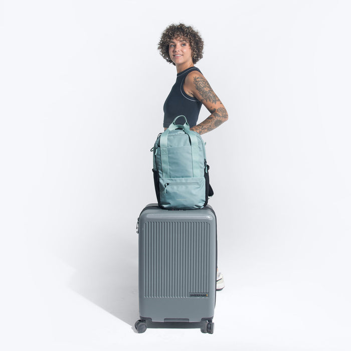 Woman holding a Sherpani Camden Surf Blue Backpack on top Sherpani Meridian Granite luggage on white background.   #color_surf-blue