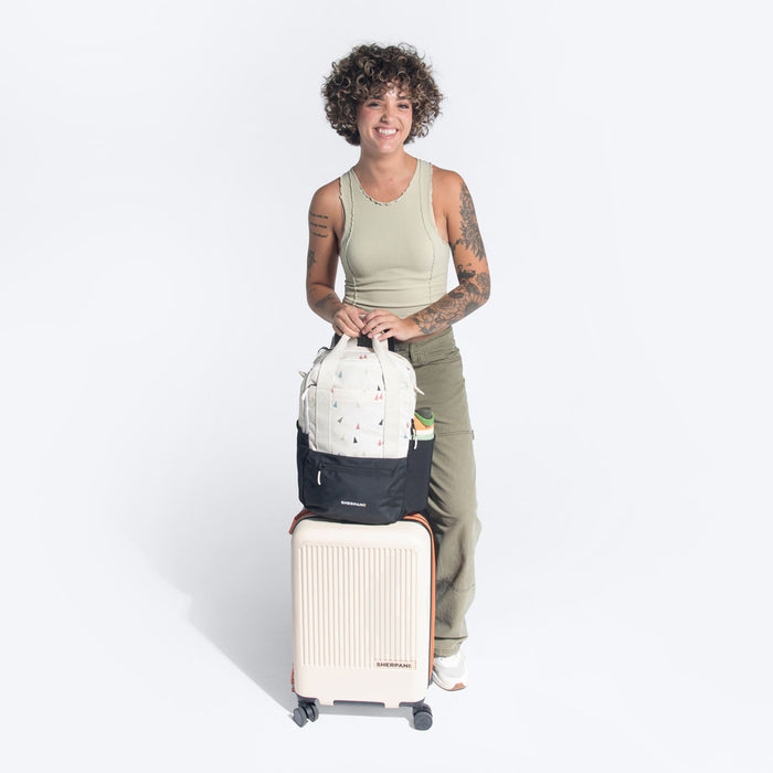 Woman holding a Sherpani Camden Terrazzo Print Backpack on top Sherpani Meridian Chai Beige luggage on white background.   #color_terrazzo-print