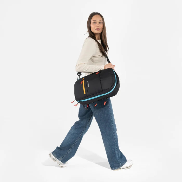 A woman wearing a Sherpani Camden Chromatic Black Ultimate Backpack with crossbody strap against white background. #color_chromatic-black