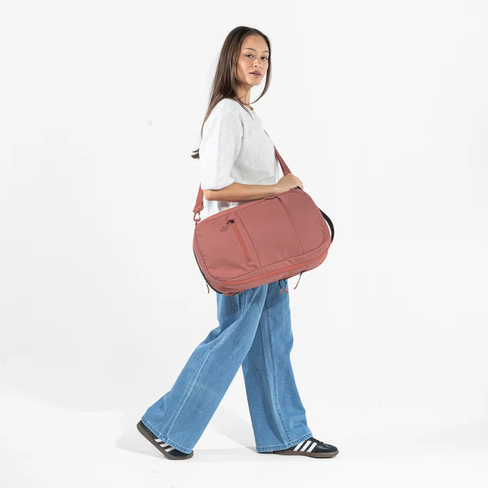 A woman wearing a Sherpani Camden Desert Pink Ultimate Backpack with crossbody strap against white background #color_desert-pink