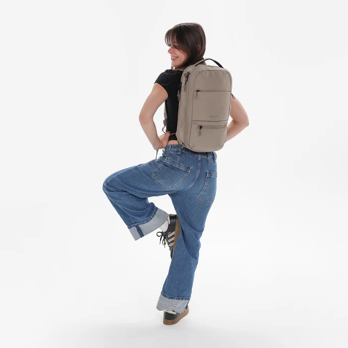 A woman wearing a Sherpani Camden Dune Ultimate Backpack on her back against white background.  #color_dune-tan
