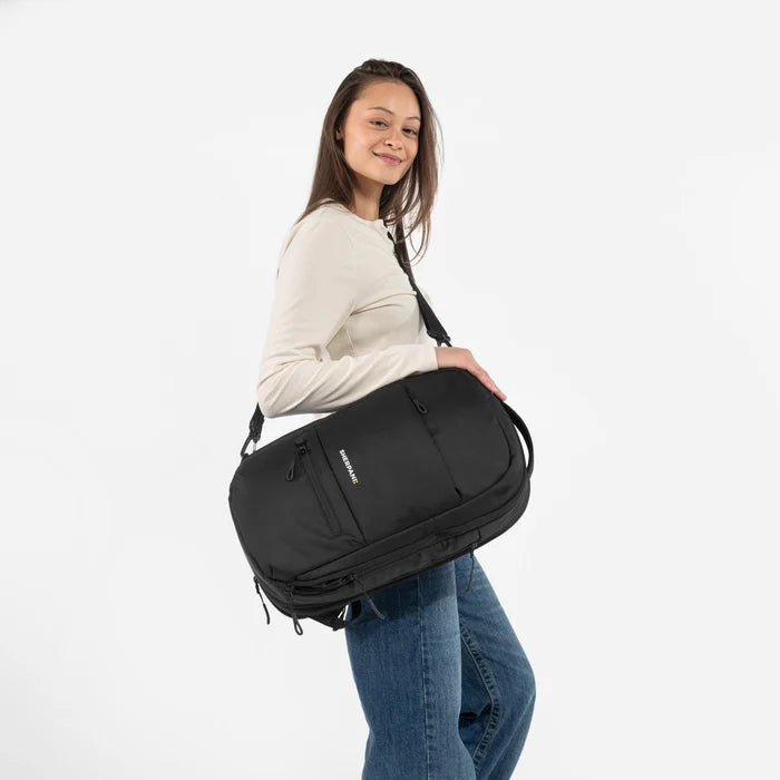 A woman wearing a Sherpani Camden Raven Black Ultimate Backpack with crossbody strap against white background #color_raven-black