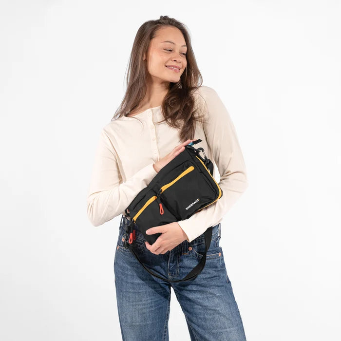 A woman holding a Sherpani Ceara Chromatic Black Expandable Crossbody against a white background.  #color_chromatic-black