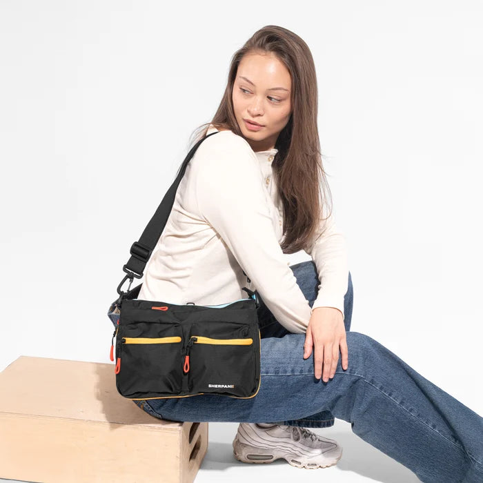A woman wearing a Sherpani Ceara Chromatic Black Expandable Crossbody while sitting against a white background. #color_chromatic-black