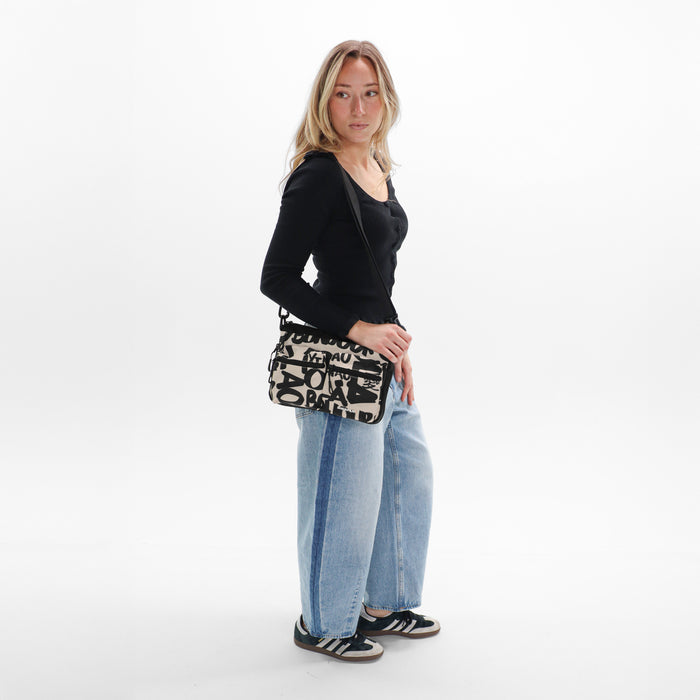 A woman wearing a Sherpani Ceara Ciao Expandable Crossbody on her shoulder against a white background.   #color_ciao-print
