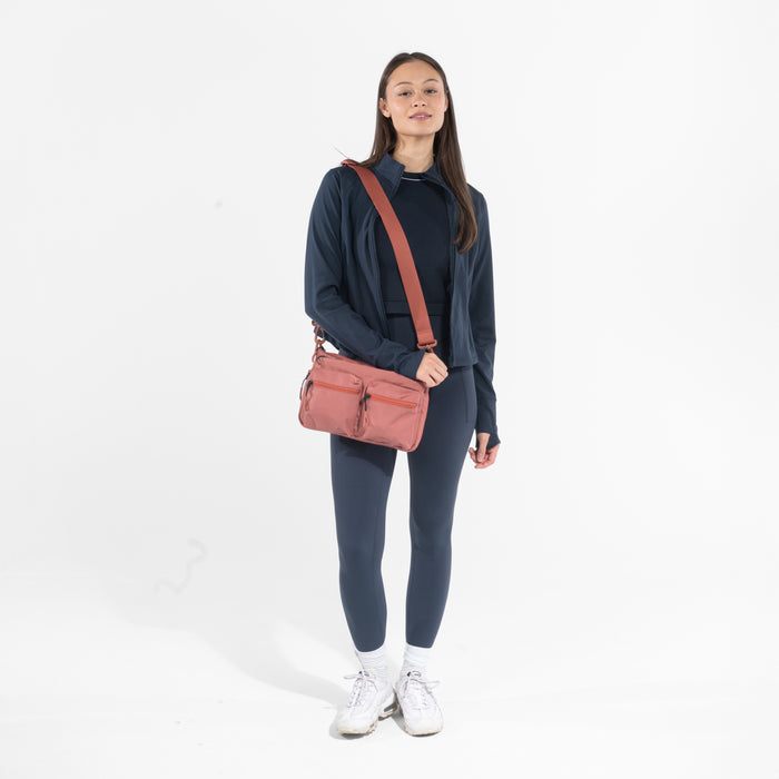 A woman wearing a Sherpani Ceara Desert Pink Expandable Crossbody on her shoulder against a white background.  #color_desert-pink