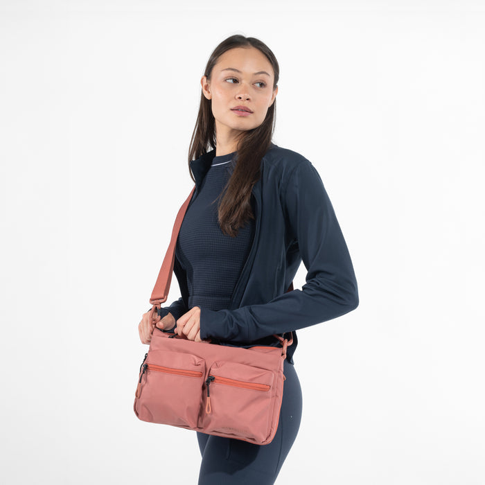A woman wearing a Sherpani Ceara Desert Pink Expandable Crossbody on her shoulder against a white background.   #color_desert-pink