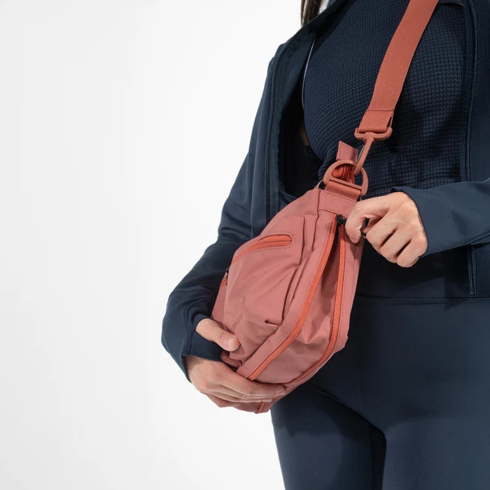 A woman unzipping the expandable pocket on a Sherpani Ceara Desert Pink Expandable Crossbody against a white background. #color_desert-pink