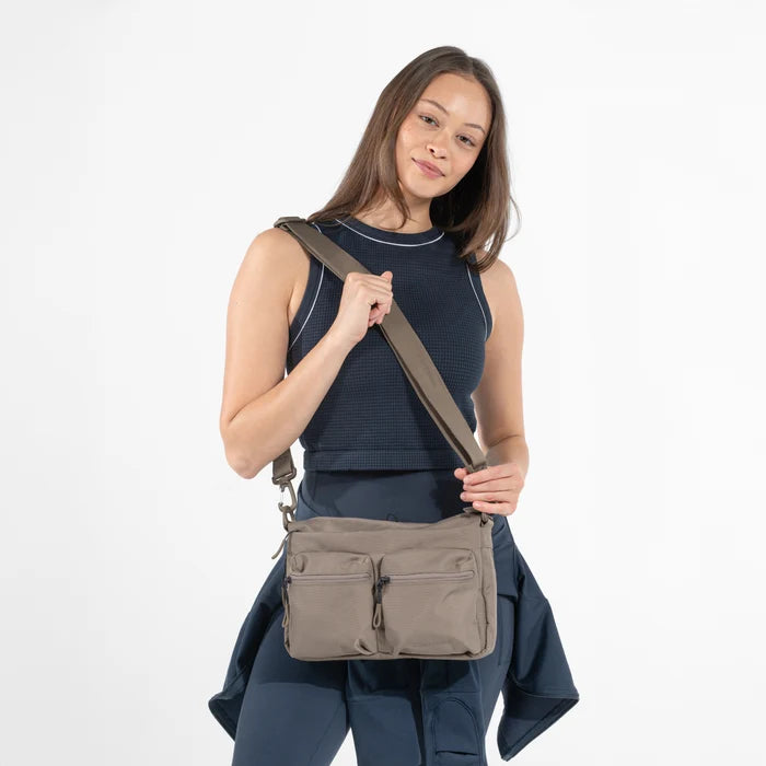 A woman wearing a Sherpani Ceara Dune Expandable Crossbody while sitting against a white background.  #color_dune-tan
