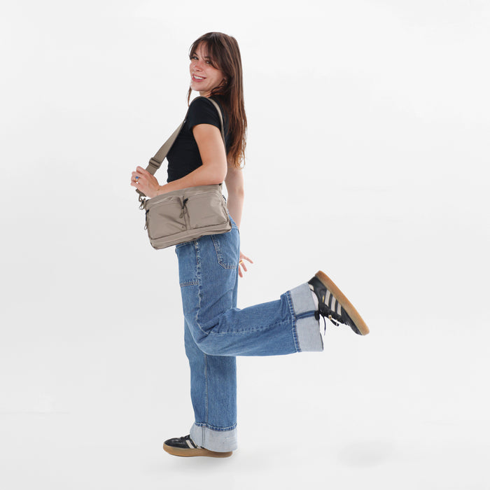 A woman wearing a Sherpani Ceara Dune  Expandable Crossbody on her shoulder against a white background.   #color_dune-tan
