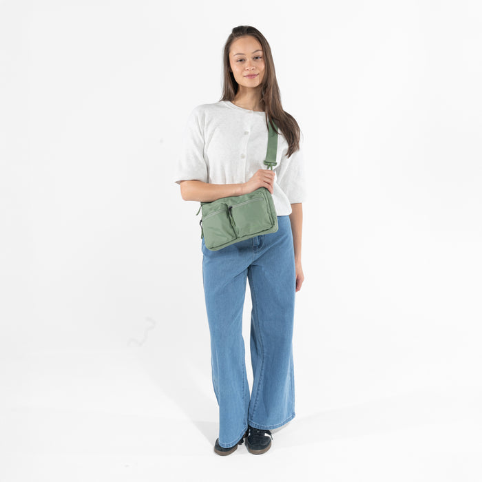 A woman wearing a Sherpani Ceara Olivine Expandable Crossbody on her shoulder against a white background.   #color_olivine-green