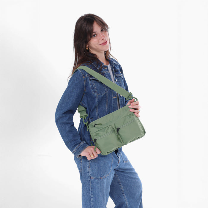A woman wearing a Sherpani Ceara Olivine Expandable Crossbody on her shoulder against a white background.   #color_olivine-green