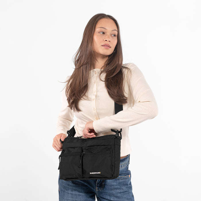 A woman wearing a Sherpani Ceara Raven Black Expandable Crossbody on her shoulder against a white background. #color_raven-black