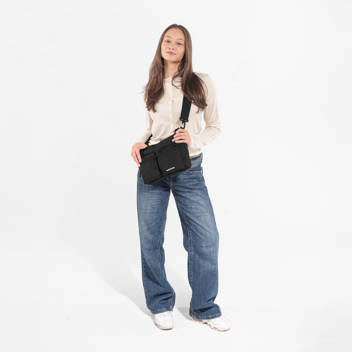 A woman wearing a Sherpani Ceara Raven Black Expandable Crossbody on her shoulder against a white background.   #color_raven-black