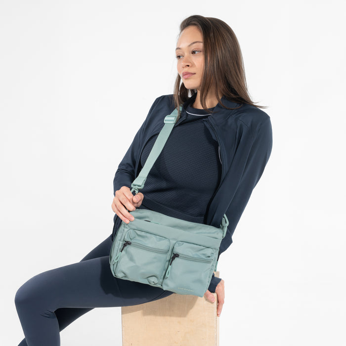 A woman wearing a Sherpani Ceara Surf Blue Expandable Crossbody while sitting against a white background. #color_surf-blue