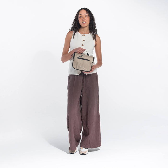 Woman holding a Sherpani Co-Pilot Straw Beige Travel Toiletry Case against white backdrop. #color_straw-beige