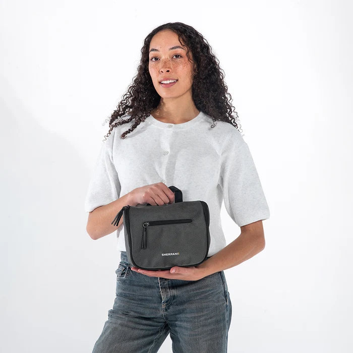 Woman holding a Sherpani Co-Pilot Granite Travel Toiletry Case against white backdrop. #color_granite-grey