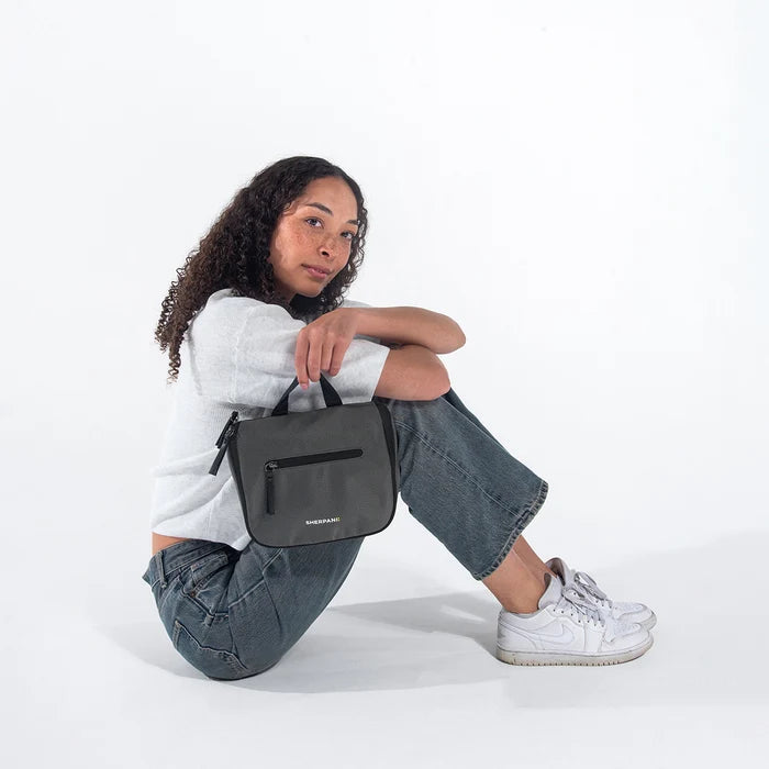 Woman holding a Sherpani Co-Pilot Granite Travel Toiletry Case against white backdrop. #color_granite-grey