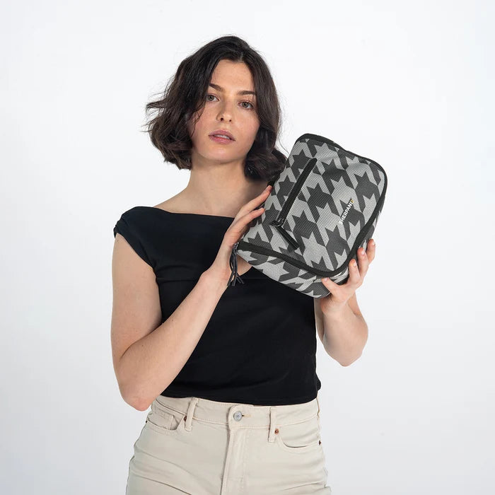 Woman holding a Sherpani Co-Pilot Houndstooth Travel Toiletry Case against white backdrop. #color_houndstooth-print