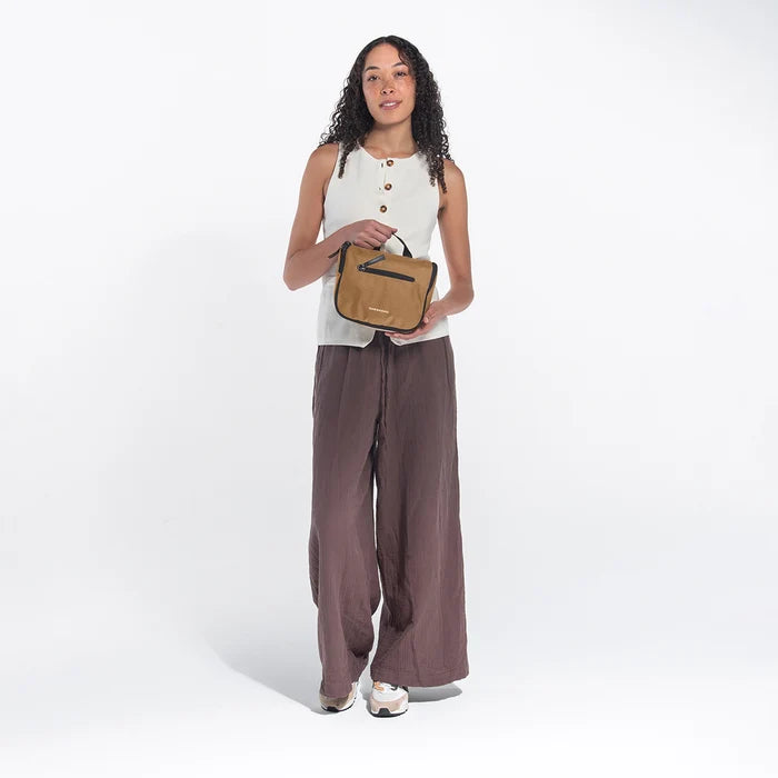 Woman holding a Sherpani Co-Pilot Sand Travel Toiletry Case against white backdrop. #color_sand-tan