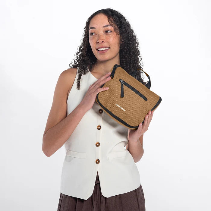 Woman holding a Sherpani Co-Pilot Sand Travel Toiletry Case against white backdrop. #color_sand-tan