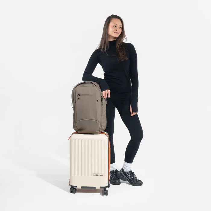 A woman holding a Sherpani Crew Dune Travel Backpack on top of a Sherpani Meridian Luggage against a white background. #color_dune-tan
