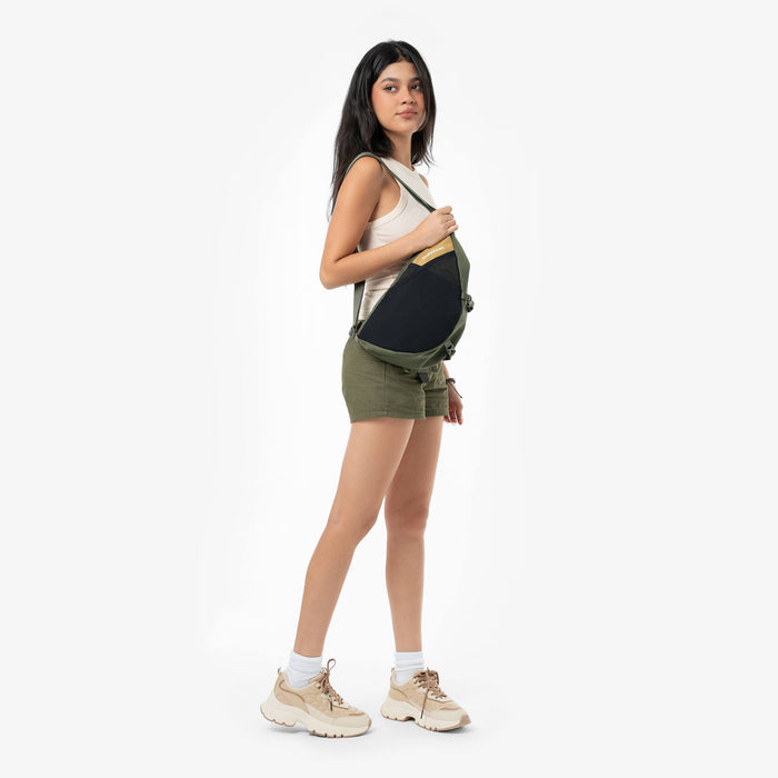 Woman wearing a Sherpani Cross Link Antelope Adventure Sling on her shoulder against white backdrop. #color_antelope