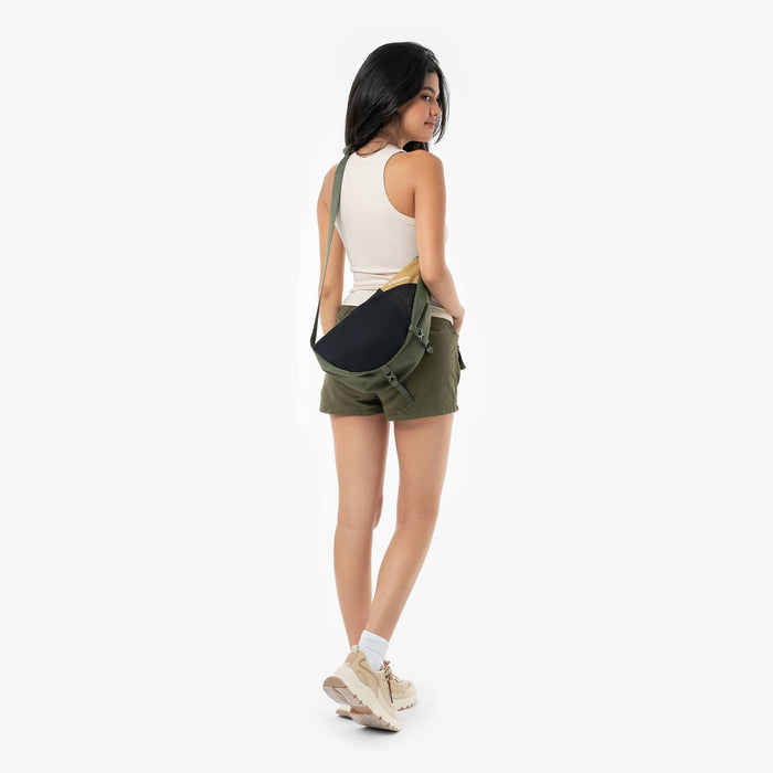 Woman wearing a Sherpani Cross Link Antelope Adventure Sling on her shoulder against white backdrop. #color_antelope
