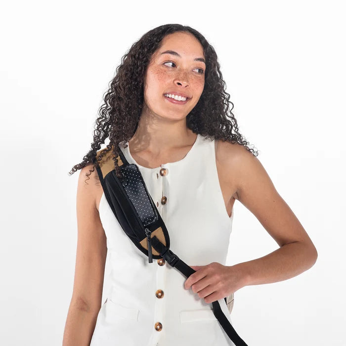 Woman wearing a Sherpani Esprit Sand Anti-theft Sling on her back with a phone in strap pocket against white backdrop. #color_sand-tan