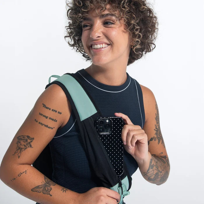 A Woman placing a phone into the stap pocket of a Sherpani Esprit Surf Blue Sling against white backdrop. #color_surf-blue