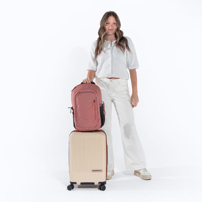 Woman leaning on a Sherpani Getaway Desert Pink travel Backpack on top Sherpani Meridian Chai Beige luggage on white backdrop. #color_desert-pink