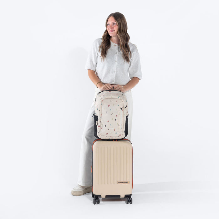 Woman leaning on a Sherpani Getaway Terrazzo Print travel Backpack on top Sherpani Meridian Chai Beige luggage on white backdrop. #color_terrazzo-print