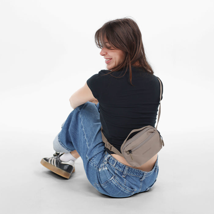 Woman wearing a Sherpani Hyk Dune Hip Pack across her back against white backdrop. #color_dune-tan