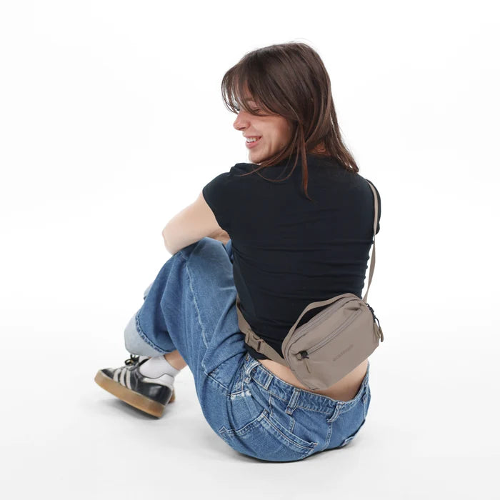 Woman wearing a Sherpani Hyk Dune Hip Pack across her back against white backdrop. #color_dune-tan