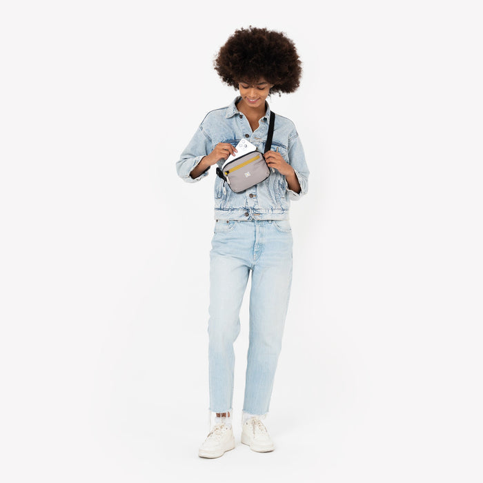 Woman placing a phone into a Sherpani Hyk Wasabi Stone Hip Pack across her body against white backdrop. #color_wasabi-stone-heritage