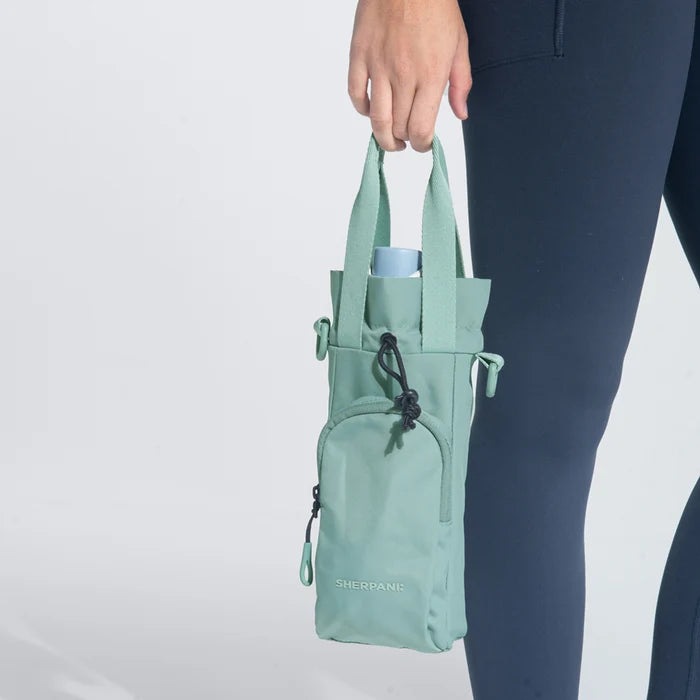 Woman holding a Sherpani Izzie Surf Blue Water Bottle Crossbody by handheld straps on white background.  #color_surf-blue