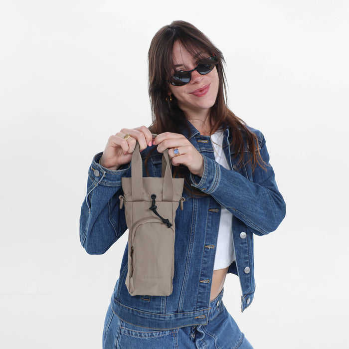 Woman holding a Sherpani Izzie Dune Water Bottle Crossbody by handheld straps on white background.   #color_dune-tan
