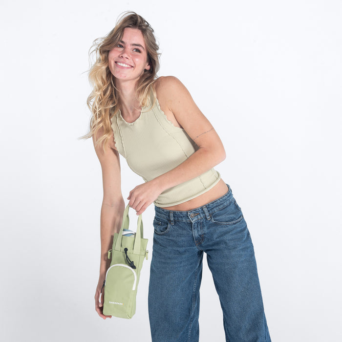 Woman holding a Sherpani Izzie Palm Green Water Bottle Crossbody by handheld straps on white background.  #color_palm-green