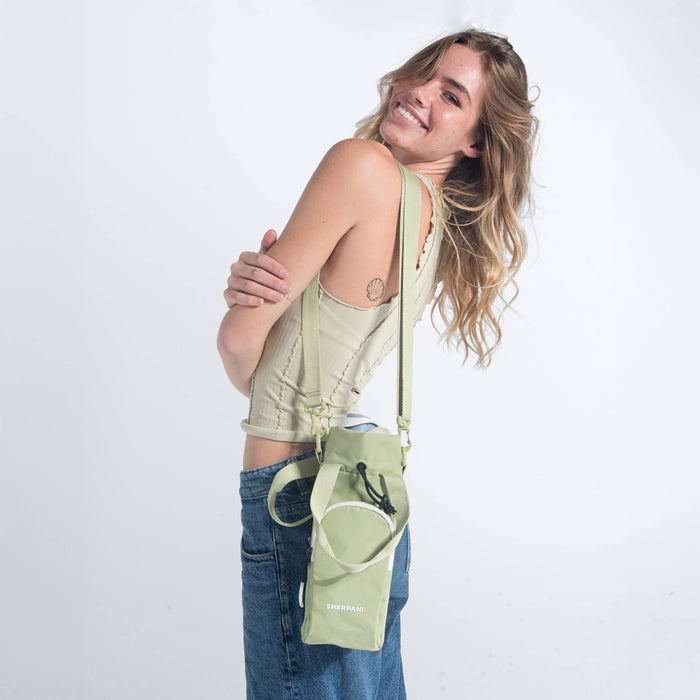 Woman wearing a Sherpani Izzie Palm Green Water Bottle Crossbody by handheld straps on white background.  #color_palm-green