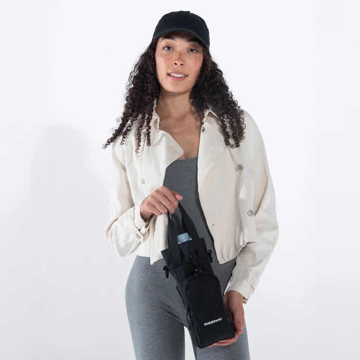 Woman holding a Sherpani Izzie Raven Black Water Bottle Crossbody by handheld straps on white background.  #color_raven-black