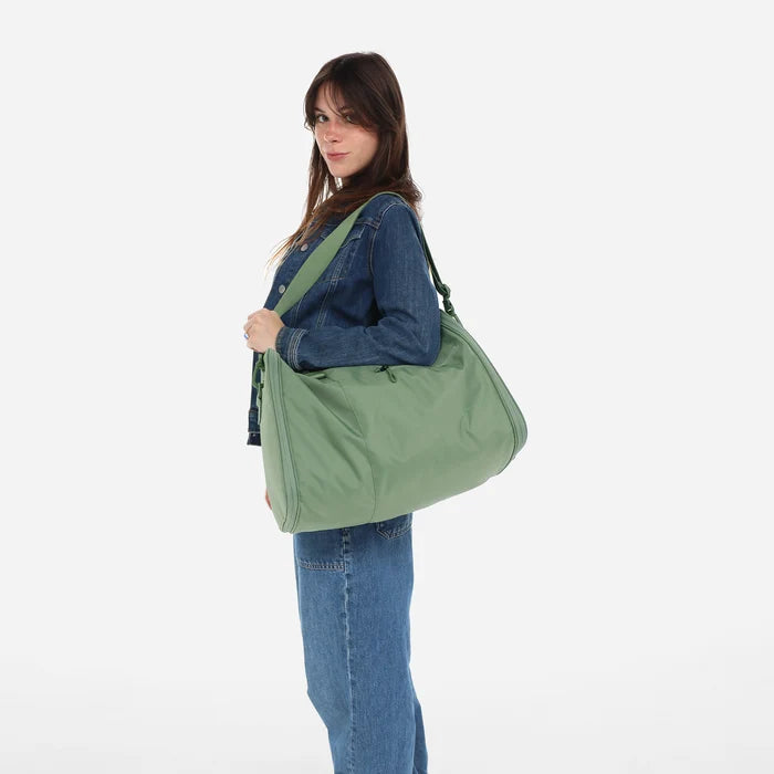 Woman wearing a Sherpani Jaunt Olivine Green Garment Duffle by the crossbody strap against white backdrop #color_olivine-green
