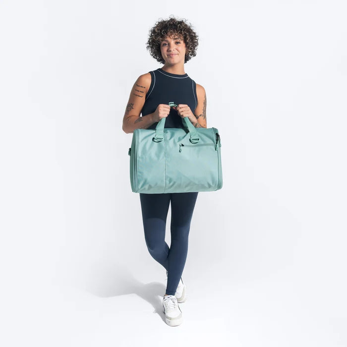 Woman holding a Sherpani Jaunt Surf Blue Garment Duffle by top straps against white backdrop #color_surf-blue