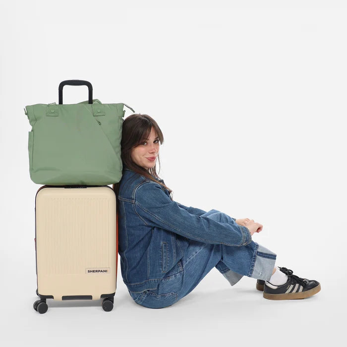 A woman sitting next to a Sherpani Olivine print Travel Tote Bag on top of A Sherpani Meridian Luggage.  #color_olivine-green