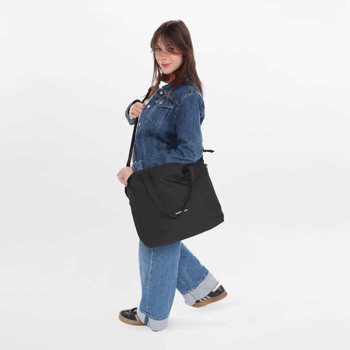 A woman wearing a Sherpani Jetsetter Raven Black Travel Tote Bag by the crossbody strap on white backdrop.  #color_raven-black