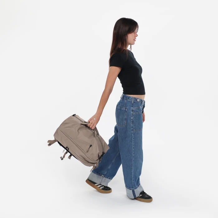 Woman holding a Sherpani Juno Dune Duffel Backpack by duffle straps against white backdrop.  #color_dune-tan