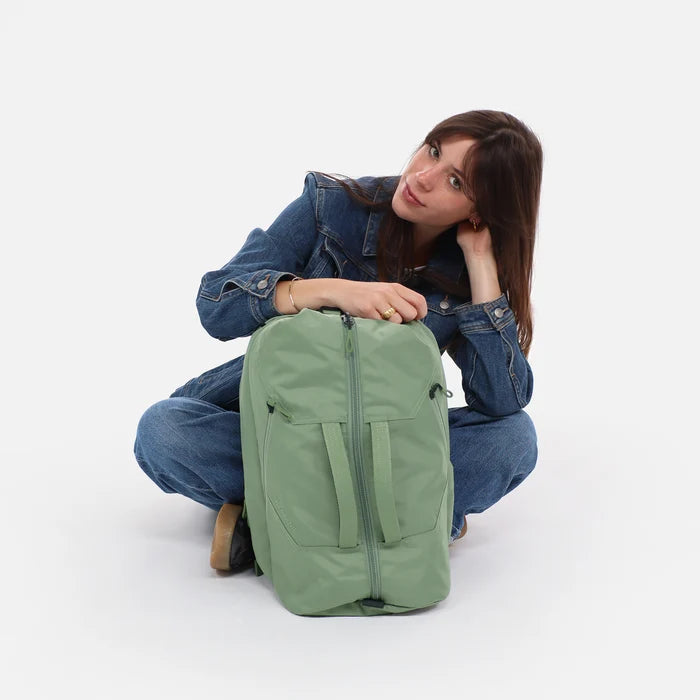 Woman sitting next to a Sherpani Juno Olivine Duffel Backpack against white backdrop. #color_olivine-green